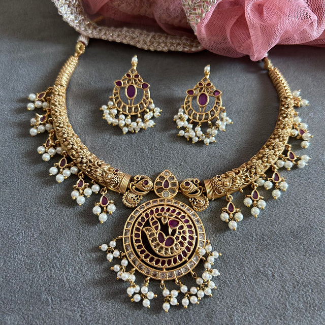 Gold earrings with gemstones on a gray surface RAKSHANA Elegant Temple Jewellery Set with INTRICATE PEACOCK DESIGN | ANTIQUE FINISH | Gold Plated RED & WHITE Stones | AD Stone Necklace set