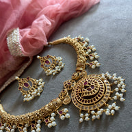 Gold necklace with pearls and red stones on a gray surface with pink fabric. RAKSHANA Elegant Temple Jewellery Set with INTRICATE PEACOCK DESIGN | ANTIQUE FINISH | Gold Plated RED & WHITE Stones | AD Stone Necklace set