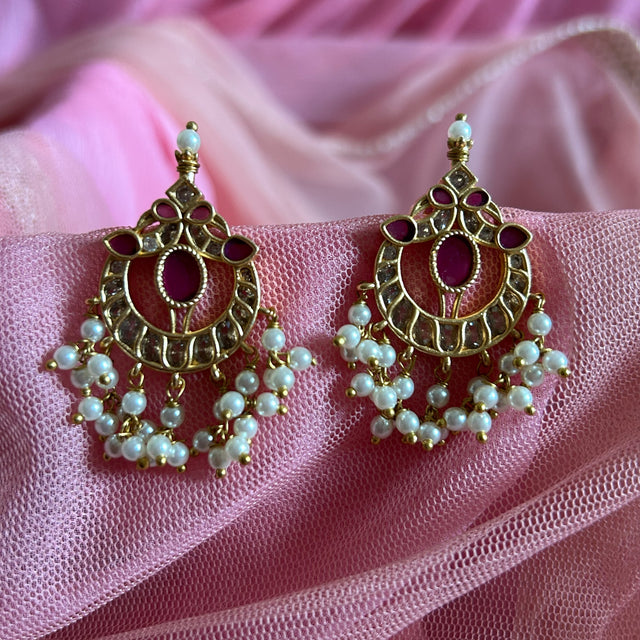 Ornate gold earrings with red stones and pearl-like beads on a pink fabric background RAKSHANA Elegant Temple Jewellery Set with INTRICATE PEACOCK DESIGN | ANTIQUE FINISH | Gold Plated RED & WHITE Stones | AD Stone Necklace set