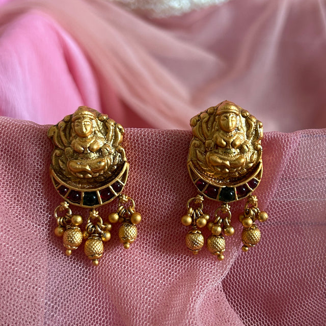 Gold earrings with intricate LAKSHMI TEMPLE designs on a pink fabric background