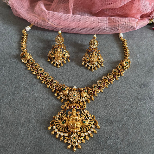 Gold necklace and earrings set on a gray surface with pink fabric in the background Gold necklace set with matching earrings on a gray surface IMITATION ARTIFICIAL ART FASHION JEWELLERY PINK STONES AD ANTIQUE PEACOCK DESIGN GOLD PLATED 