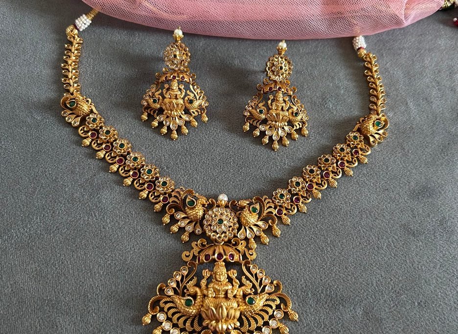 Gold necklace and earrings set on a gray surface with pink fabric in the background Gold necklace set with matching earrings on a gray surface IMITATION ARTIFICIAL ART FASHION JEWELLERY PINK STONES AD ANTIQUE PEACOCK DESIGN GOLD PLATED 